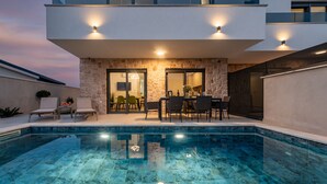 Uma área de piscina moderna com parede de pedra, mobiliário de exterior e piscina translúcida.
