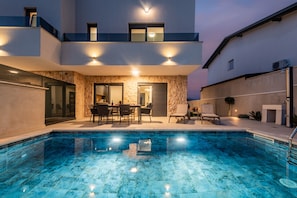 A modern pool area with a stone facade, a dining area with chairs and a table, and a clear blue pool. - Seaview Villa Sal with pool, jacuzzi and sauna (Privlaka)