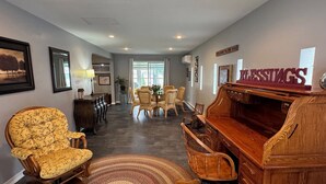 A living room with a piano, wicker chairs, a patterned armchair, and a 'WELCOME TO THE ZOO' sign. - Just Peachy! Enjoy your private lake front home. (West Branch)