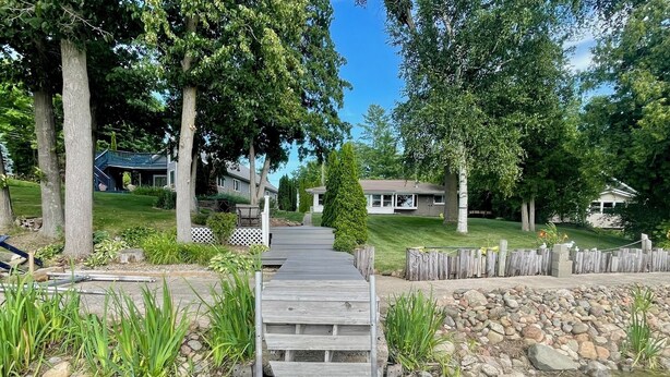 A wooden deck leading to a pond with a stone wall and trees in the background. - Just Peachy! Enjoy your private lake front home. (West Branch)