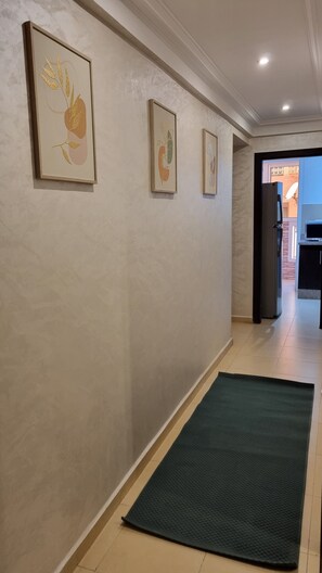 Ein Flur mit drei gerahmten Kunstwerken an der Wand, einer dunkelgrünen Matte auf dem Boden und einem Blick in den Küchenbereich im Hintergrund.