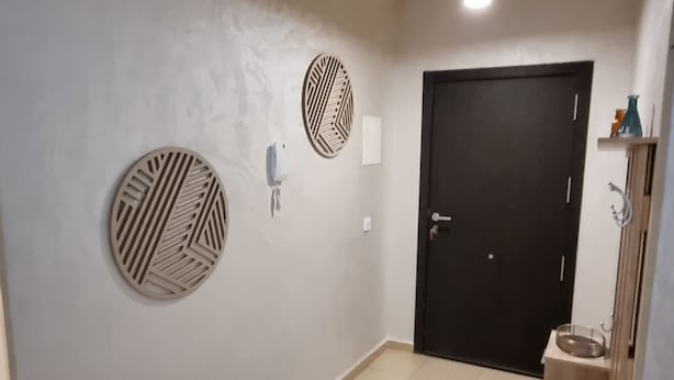 Un pasillo con suelo de baldosas, un tapete redondo, una puerta y una lámpara fijada a la pared.
