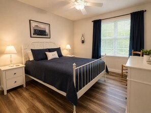 Ein Schlafzimmer mit Bett, Nachttischen, Schreibtisch, Stuhl, Deckenventilator, Fenster mit Vorhängen und einem Wandbild.