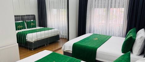 A hotel room with two beds, each with green bedspreads and white linens. There is a large mirror on one wall and a window with sheer curtains.