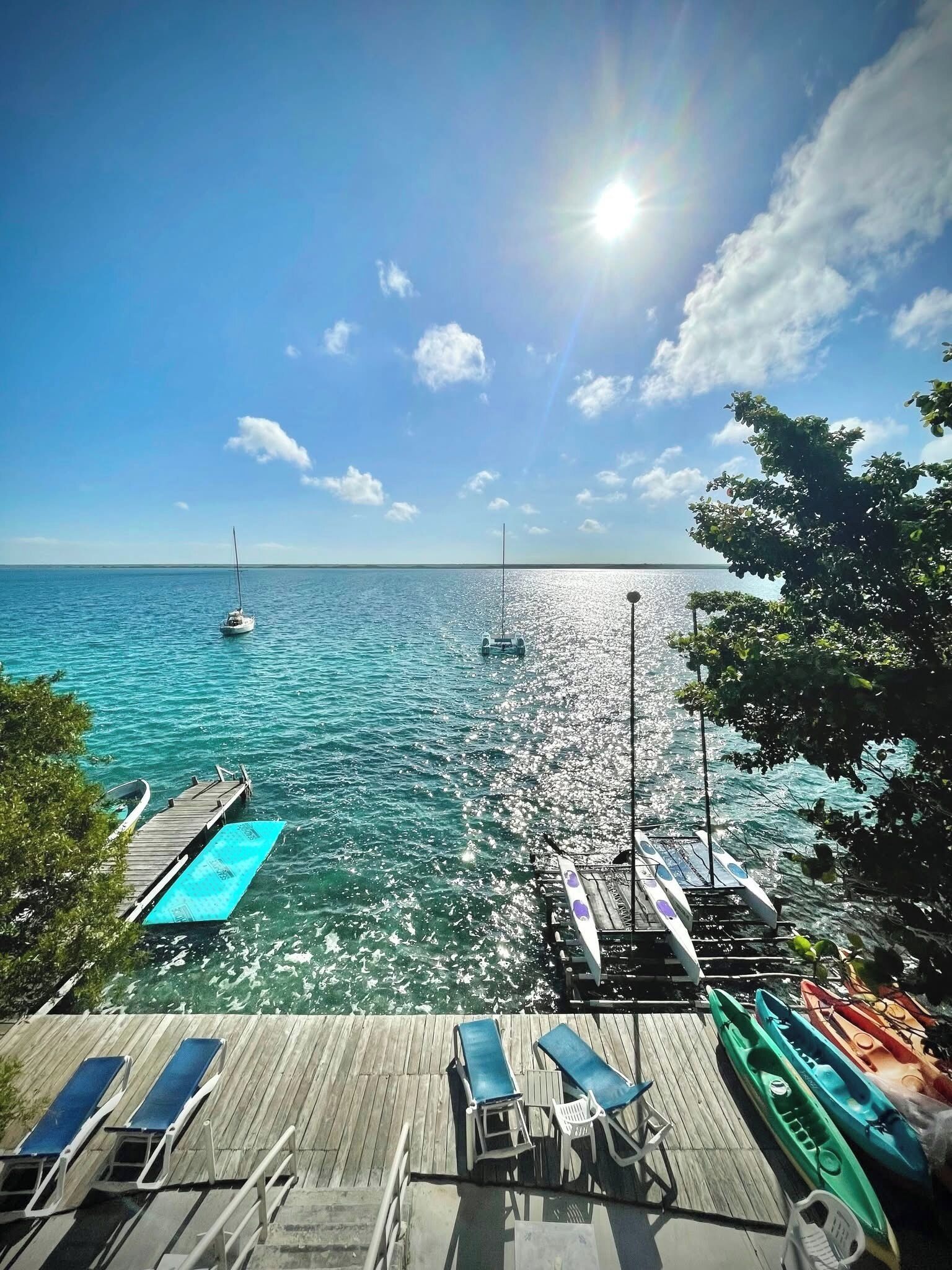 A wooden pier extending into a calm sea with a clear sky, a sailboat, and kayaks docked.