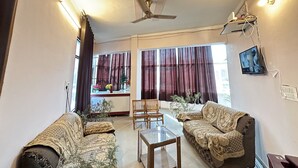 Una sala de estar con dos sofás, una mesa de centro de vidrio, un ventilador de techo y ventanas grandes con cortinas.