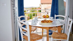 Une table ronde entourée de chaises blanches, une assiette de croissants et un panier de fruits sur un balcon agrémenté de rideaux bleus, d’où l’on peut admirer la vue sur les immeubles.
