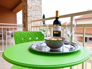 Uma mesa com uma garrafa de vinho, copos e uma tigela de biscoitos numa varanda, com um muro de pedra e telhado de azulejos ao fundo.