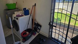 A laundry area with a washing machine, mops, brooms, and a metal gate leading to a yard. - Sun-Filled Cozy House Near Beautiful Beach (Discovery Bay, St. Ann)