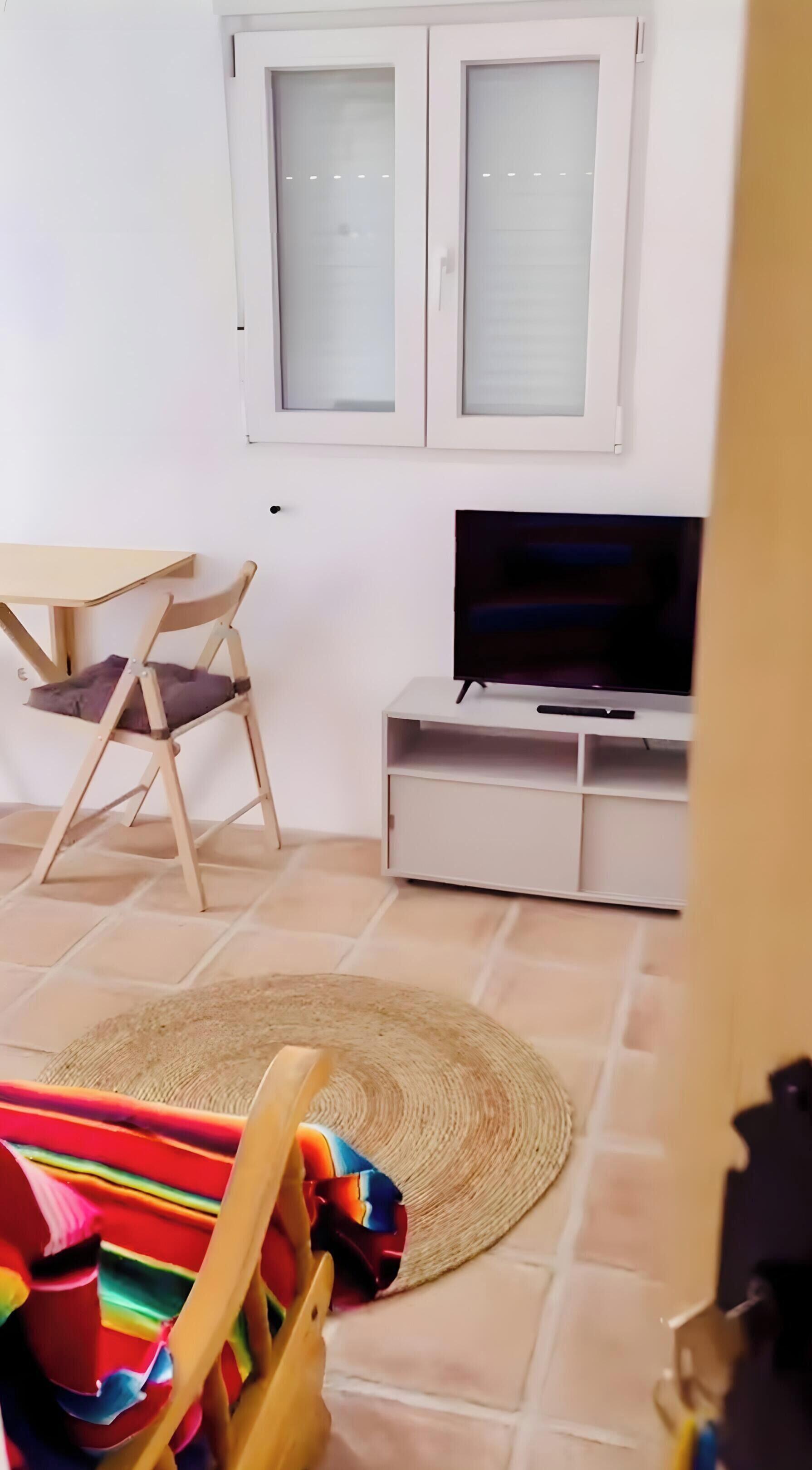 A room with a TV on a stand, a chair, a table, and a window with blinds.