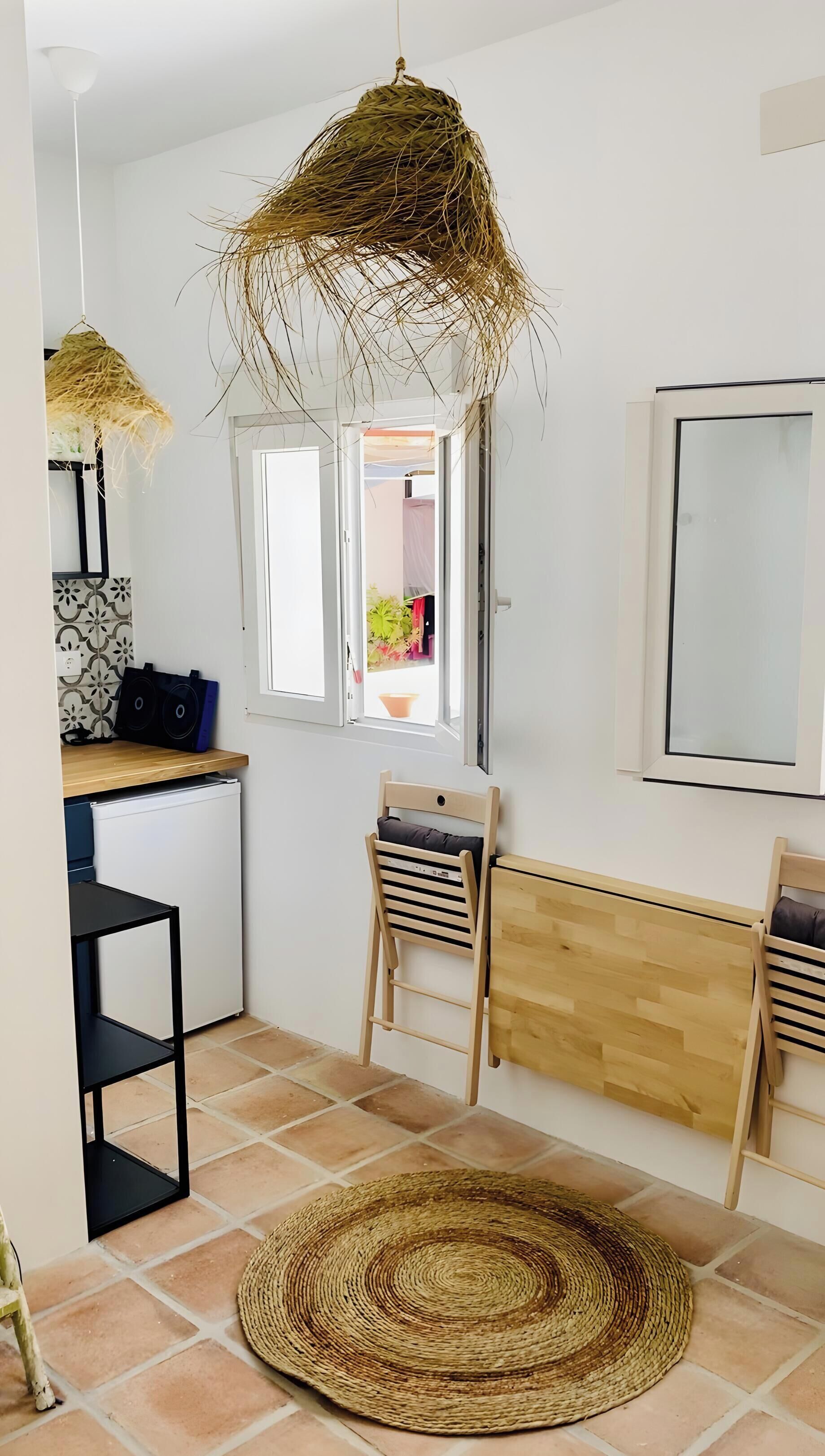 A room with a woven hanging light fixture, a window with a view of plants, a wooden desk with a chair, and a woven rug on the floor.