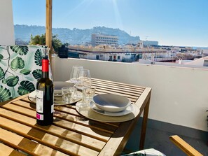 Una mesa de madera con una botella de vino, copas y un plato en un balcón con vistas a una ciudad.