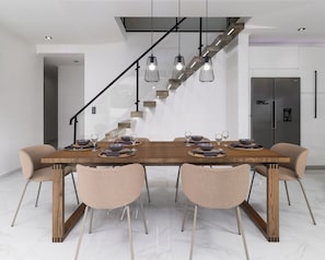 A dining area with a wooden table, six chairs, and hanging pendant lights. A staircase with a glass railing and wooden steps is visible in the background. - Villa Aurora (Ponta Delgada)