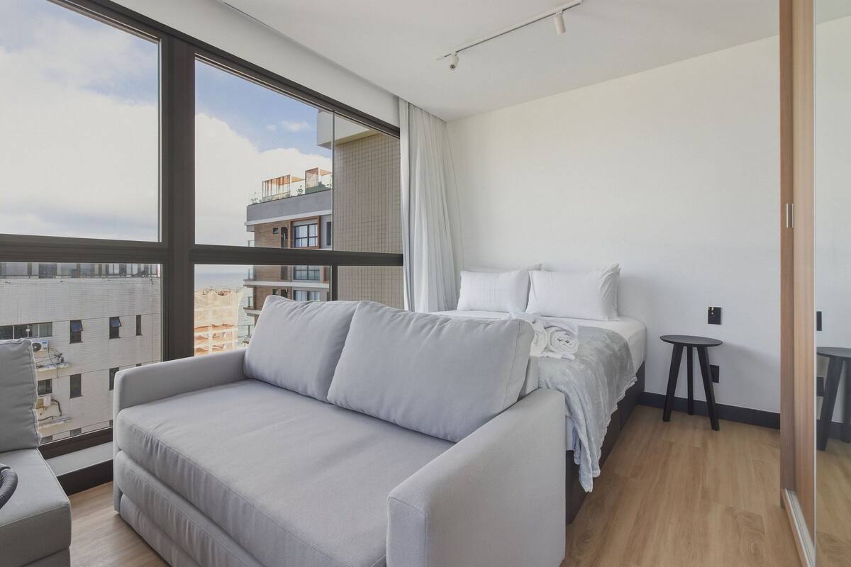 A modern hotel room with a large window, a sofa, a bed, and a small table.