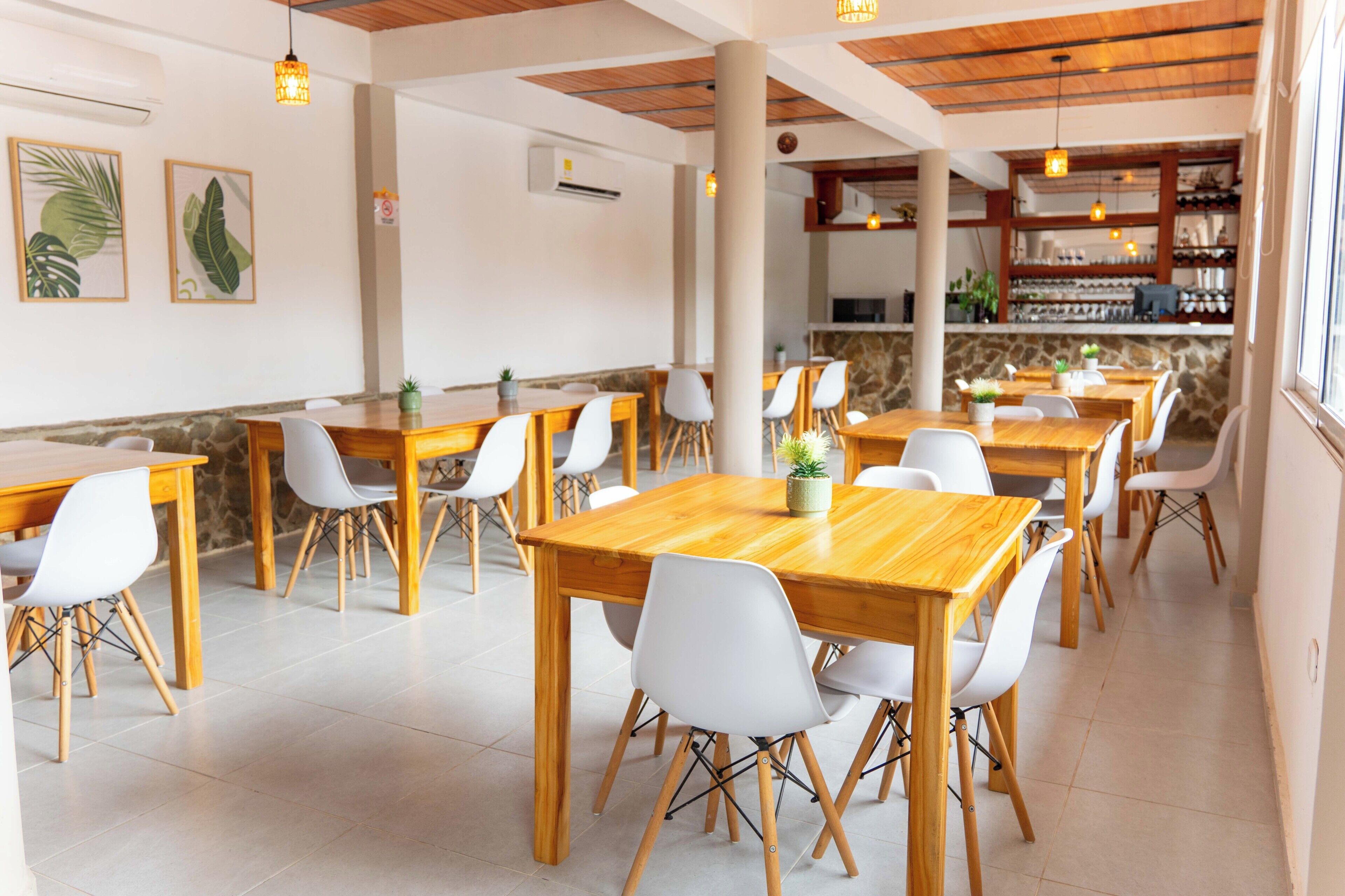 Un restaurante con varias mesas de madera y sillas blancas, una zona de bar con estantes llenos de botellas y una pared de piedra al fondo.
