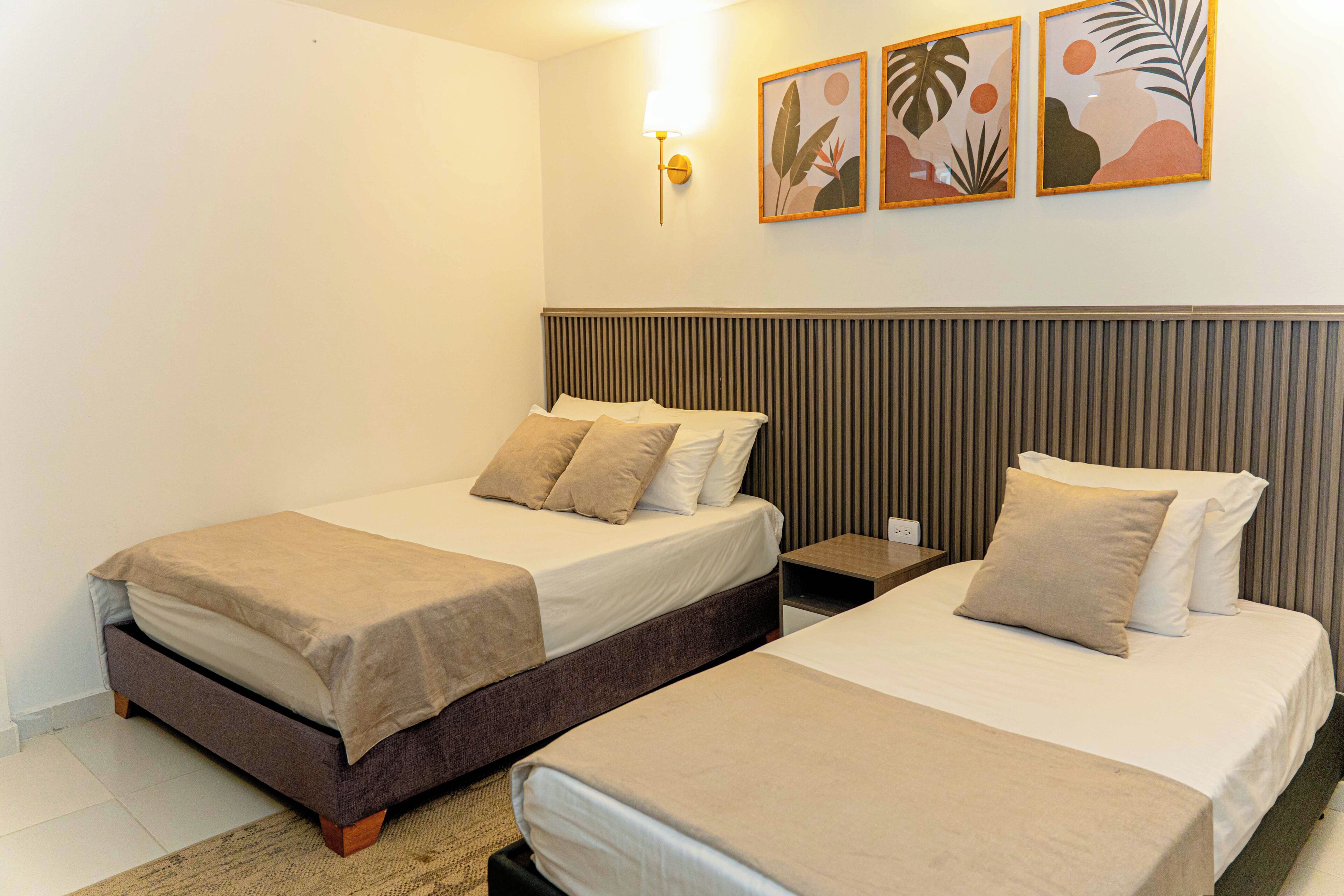 A hotel room with two beds, a nightstand, a lamp, and three framed artworks on the wall.