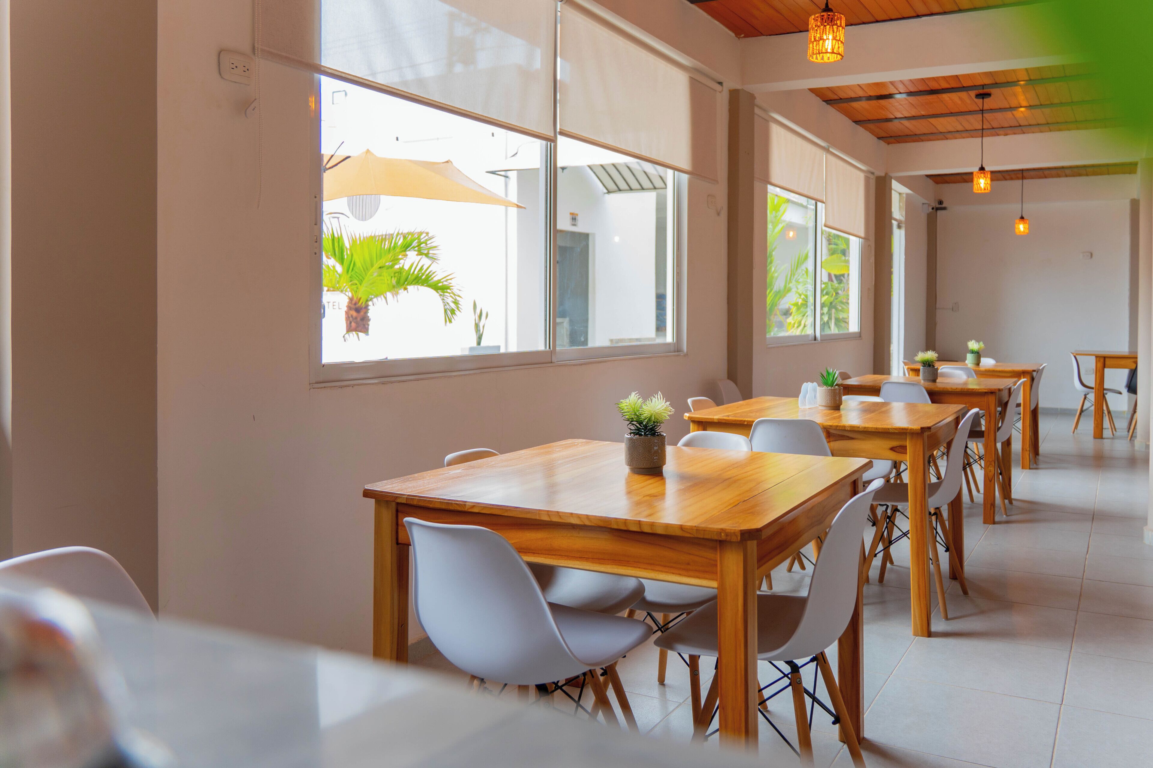 Un comedor amplio con mesas de madera y sillas blancas, ventanas grandes con persianas e iluminación colgante.