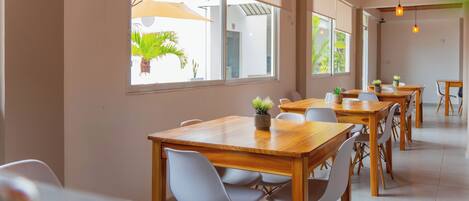 A spacious dining area with wooden tables and white chairs, large windows with blinds, and hanging pendant lights.