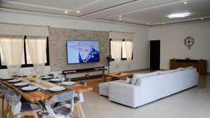 Ein modernes Wohnzimmer mit Essbereich, einem an der Wand befestigten Flachbildfernseher, einem weißen Sofa und einem Holzessttisch, gedeckt für sechs Personen.