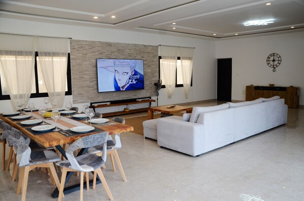 A modern living room with a dining area, a flat-screen TV mounted on the wall, a white sofa, and a wooden dining table set for six.