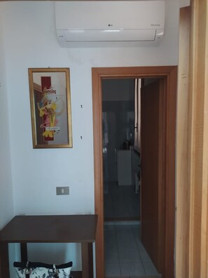 A room with a wooden table, an air conditioning unit, a framed poster, and a doorway leading to another room. - Apartment 'Bilocale Buca Del Gatto 210- Cecina' with Balcony (Mazzanta)
