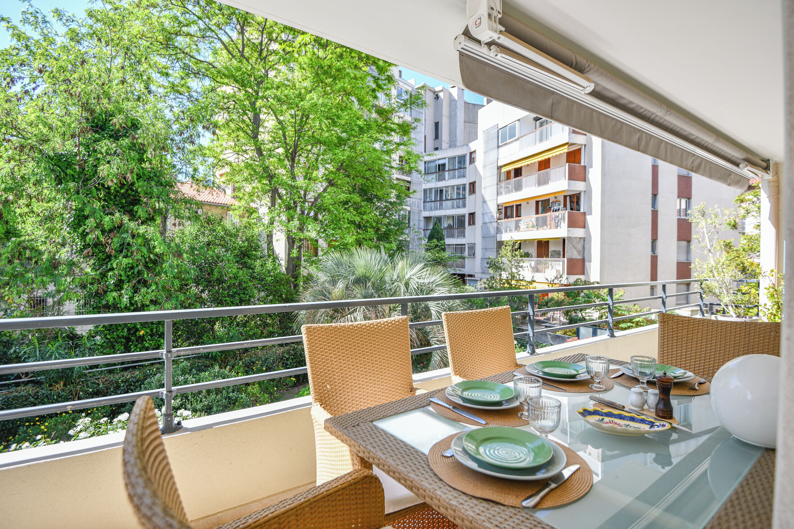 Un balcone con sedie in vimini e un tavolo in vetro apparecchiato per consumare un pasto, con vista su un rigoglioso giardino e edifici residenziali.