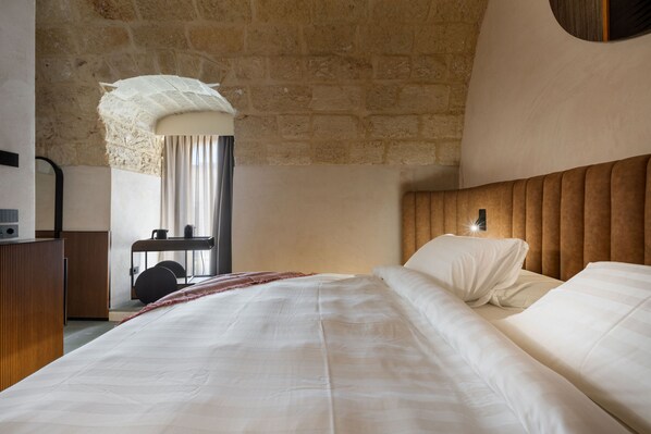 Une chambre avec un mur en pierre, un lit avec du linge de lit blanc, une tête de lit en bois et un bureau avec une chaise.