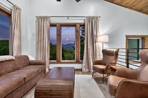 A modern living room with a sofa, armchair, and coffee table, large windows with curtains, and a view of greenery. - Modern Mt LeConte Cabin • Hot Tub + Spa Tub (Gatlinburg)