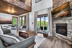 A modern living room with a fireplace, large windows, and a dining area. - Modern Mt LeConte Cabin • Hot Tub + Spa Tub (Gatlinburg)