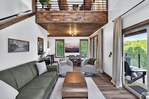 A modern living room with a wooden ceiling, a green sofa, a coffee table, and a view of greenery through large windows. - Modern Mt LeConte Cabin • Hot Tub + Spa Tub (Gatlinburg)