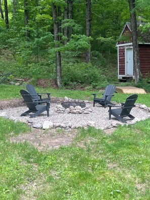 En plats med en öppen eldstad, Adirondack-stolar och ett litet förråd, belägen i en skogig miljö.