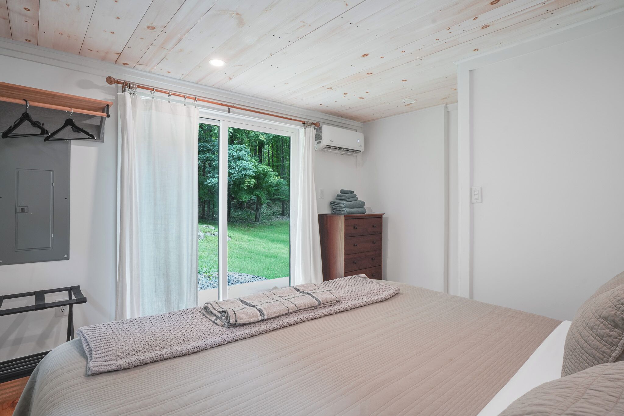 A bedroom with a large bed, a wooden dresser, a hanging coat rack, and a sliding glass door leading to a grassy outdoor area.