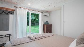 A bedroom with a large bed, a wooden dresser, a hanging coat rack, and a sliding glass door leading to a grassy outdoor area.