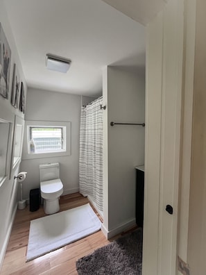 A small bathroom with a toilet, a window, a shower curtain, and a white rug on the floor.