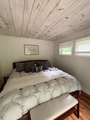 Une chambre avec un plafond en bois, un lit recouvert d’une couette blanche et une fenêtre avec des stores.