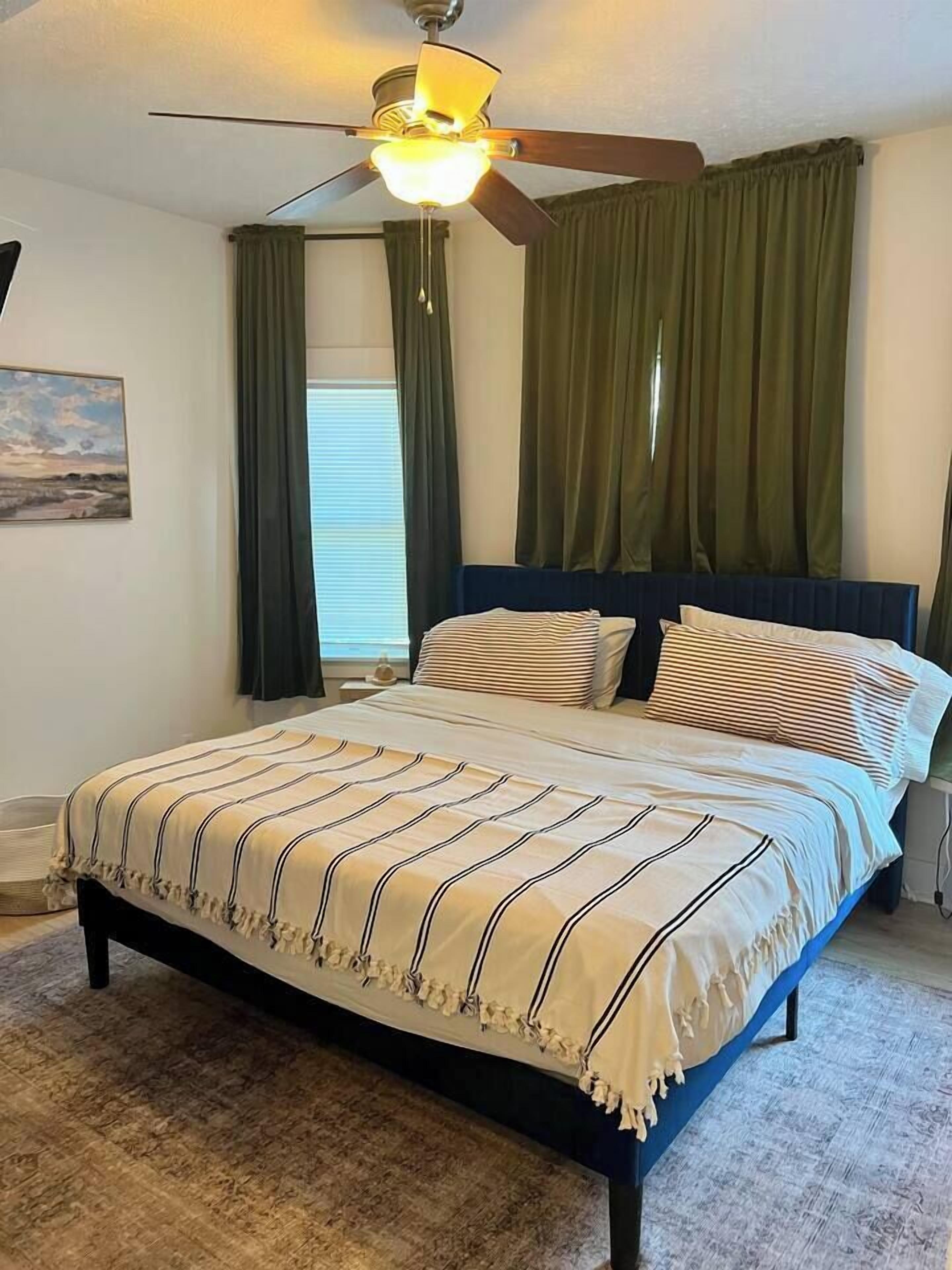 Una camera da letto con un letto, una ventola a soffitto, una finestra con tende e un quadro appeso al muro.