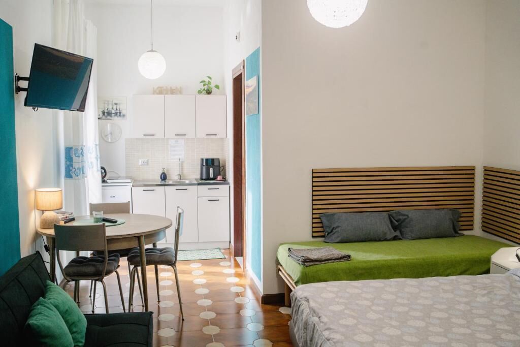 A compact room with a bed, a desk, a chair, a TV mounted on the wall, and a kitchenette.