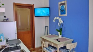 Una habitación con televisor, una puerta, una mesa con sillas, una planta pequeña y un cuadro colgado en la pared.