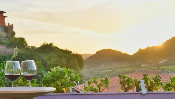 Una terraza con dos copas de vino, vista a una colina y un edificio al fondo.