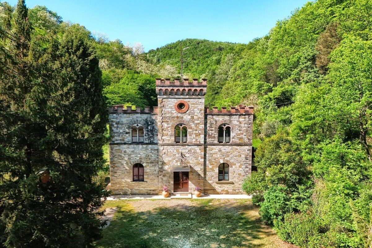 A historic stone building with a central tower, arched windows, and a paved courtyard surrounded by dense greenery.