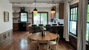 A kitchen with a round dining table, wooden chairs, pendant lights, and a view of the outdoors. - Victoria’s Chamber Fourplex | Rooftop deck and Zoo (Omaha)