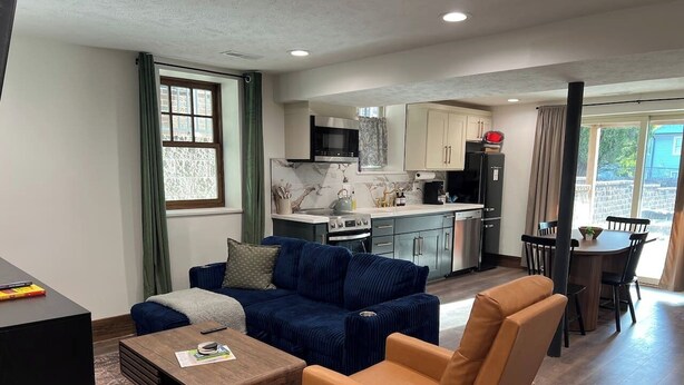A living room with a sofa, armchair, coffee table, and TV stand. There is a kitchen in the background with cabinets, a microwave, and a dining area with a table and chairs. - The Candlelight Cellar on 10th | Fourplex (Omaha)
