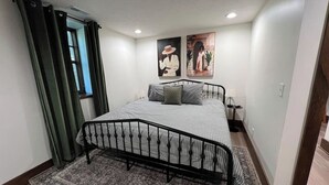 A bedroom with a bed, a rug, a window with curtains, and two wall-mounted pictures. - The Candlelight Cellar on 10th | Fourplex (Omaha)