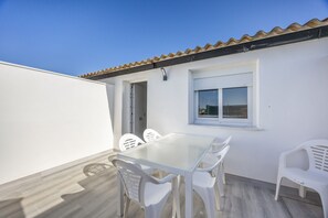 Een terras met een glazen tafel en witte plastic stoelen, een deur en een raam met zonwering.