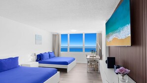 Quarto de hotel com duas camas, uma escrivaninha, uma cadeira, televisão e uma janela grande com vista para o mar.