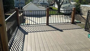 Een houten terras met een metalen hek, omgeven door woonhuizen.