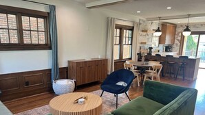 Een moderne woonkamer met een groene bank, een ronde houten salontafel en een blauwe fauteuil. Er is een eethoek met een tafel en stoelen, en een keuken op de achtergrond.