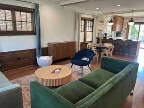 Ein modernes Wohnzimmer mit einem grünen Sofa, einem runden Holztisch und einem blauen Sessel. Es gibt eine Essecke mit Tisch und Stühlen, und im Hintergrund eine Küche.