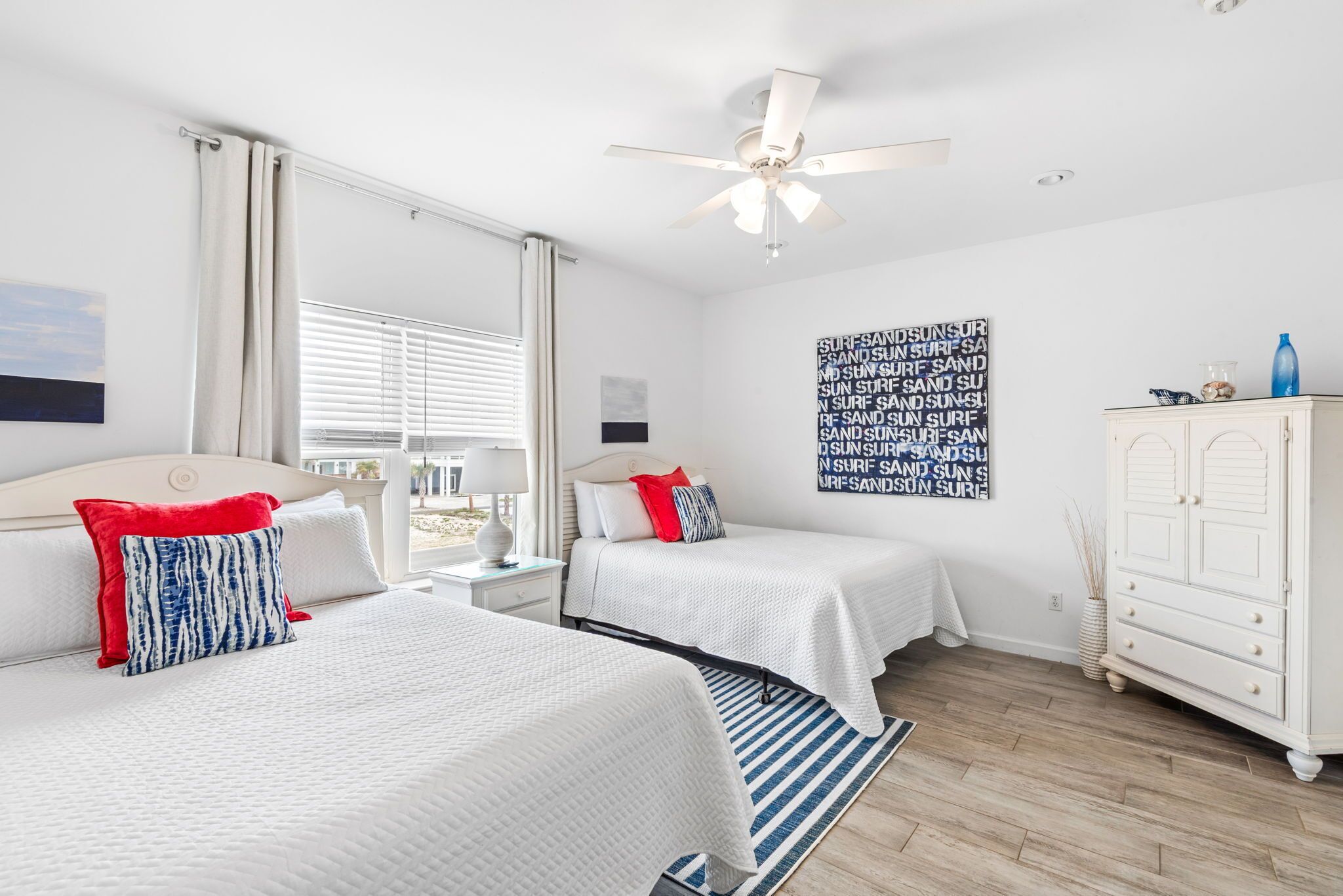 A hotel room with two beds, a ceiling fan, a large window with blinds, a wall art piece, and a white dresser.