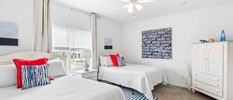 A hotel room with two beds, a ceiling fan, a large window with blinds, a wall art piece, and a white dresser.